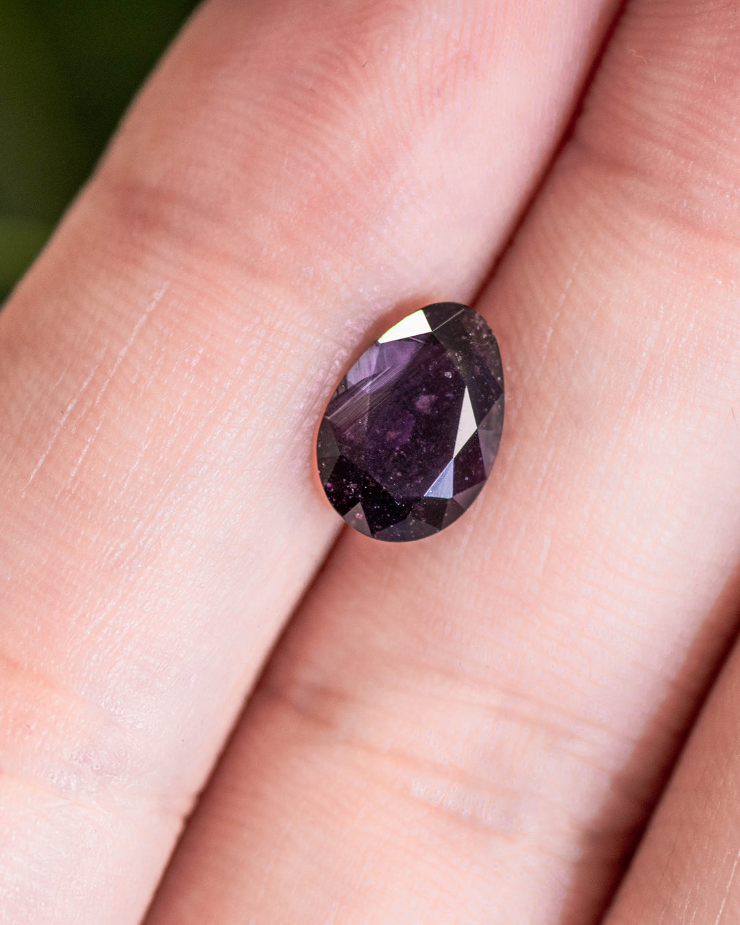 1.98 carat purple sapphire placed on fingertip under natural light, revealing rich plum undertones and precise faceting in its modified tear drop cut, untreated gemstone from Umba River, Tanzania.