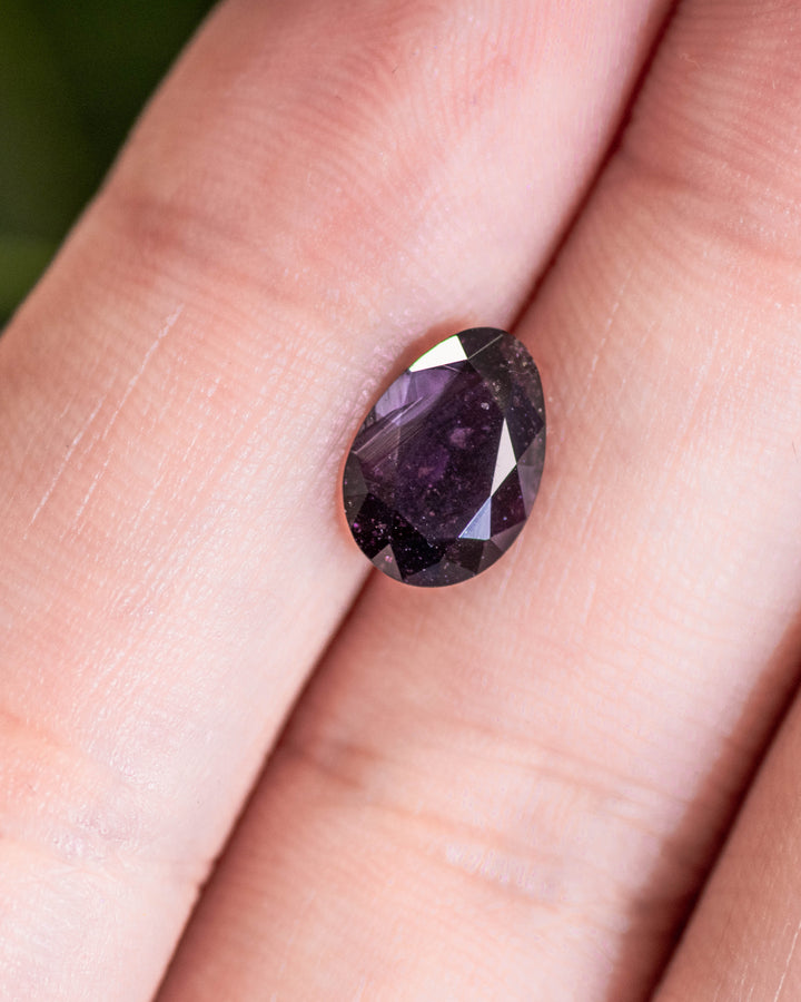 1.98 carat purple sapphire placed on fingertip under natural light, revealing rich plum undertones and precise faceting in its modified tear drop cut, untreated gemstone from Umba River, Tanzania.