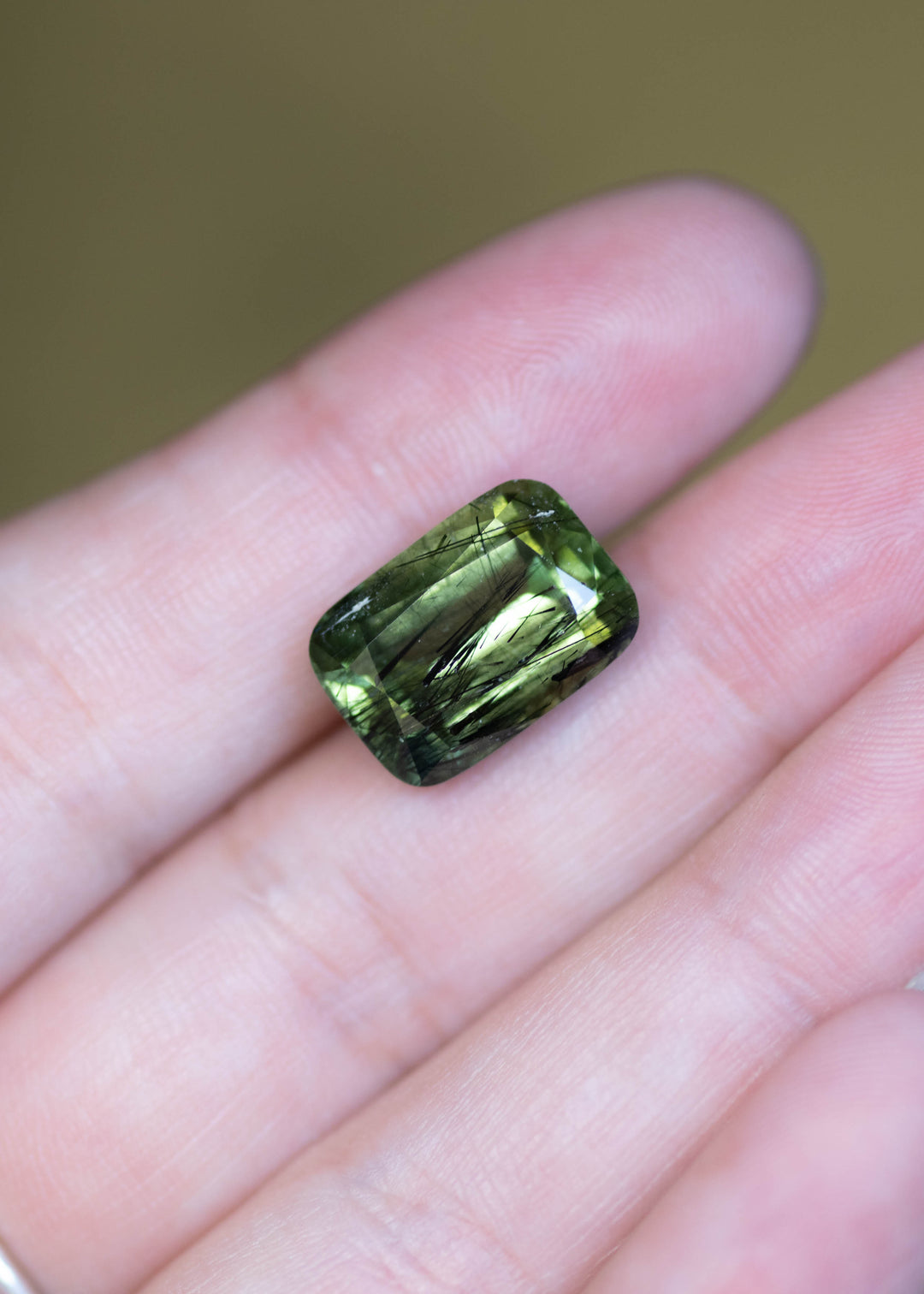 Untreated 10.89 carat forest green peridot from Pakistan shown on fingertips for scale, displaying a modified radiant cut and rich natural color.