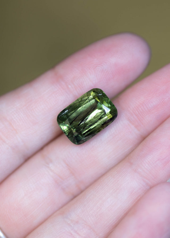 Untreated 10.89 carat forest green peridot from Pakistan shown on fingertips for scale, displaying a modified radiant cut and rich natural color.