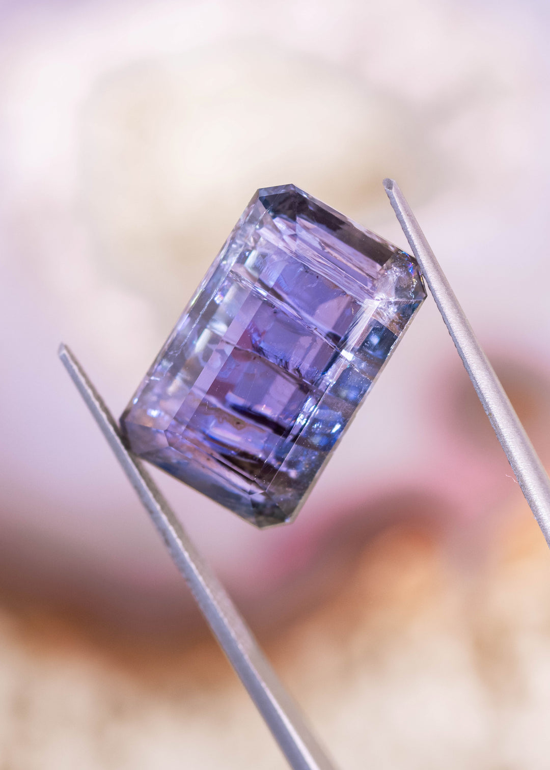 18.04 carat Tanzanite gemstone displaying teal, blue, and violet color zoning in a custom octagonal step cut, photographed in tweezers under soft light to show clarity and facet structure.