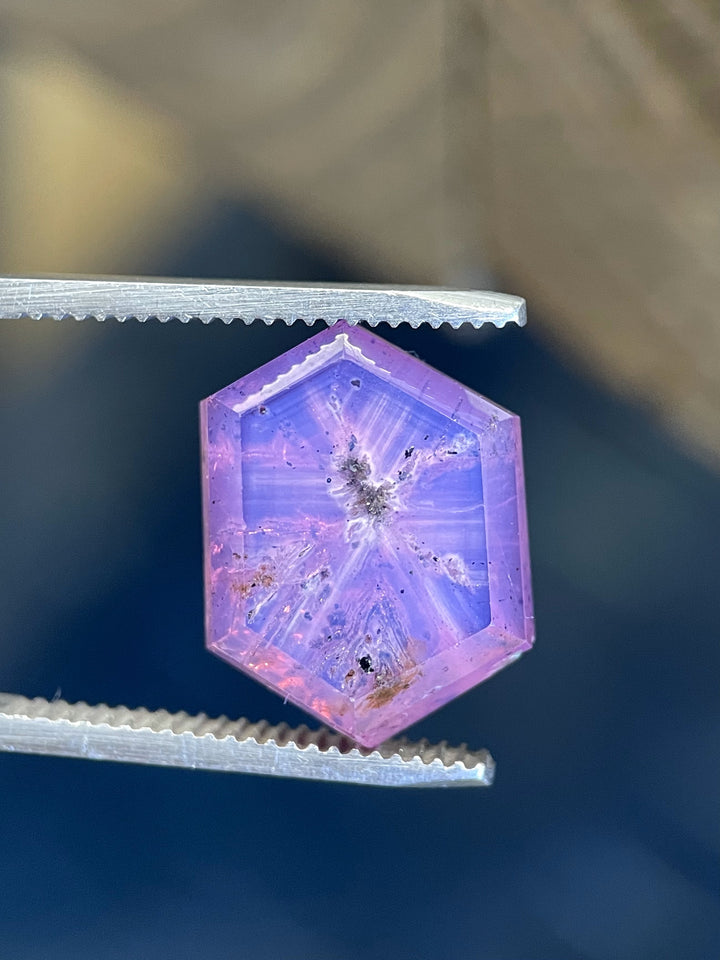 A 19.62-carat pink and purple sapphire in a hexagon portrait cut, delicately held on a fingertip, showcasing its raw beauty and intricate inclusions.
