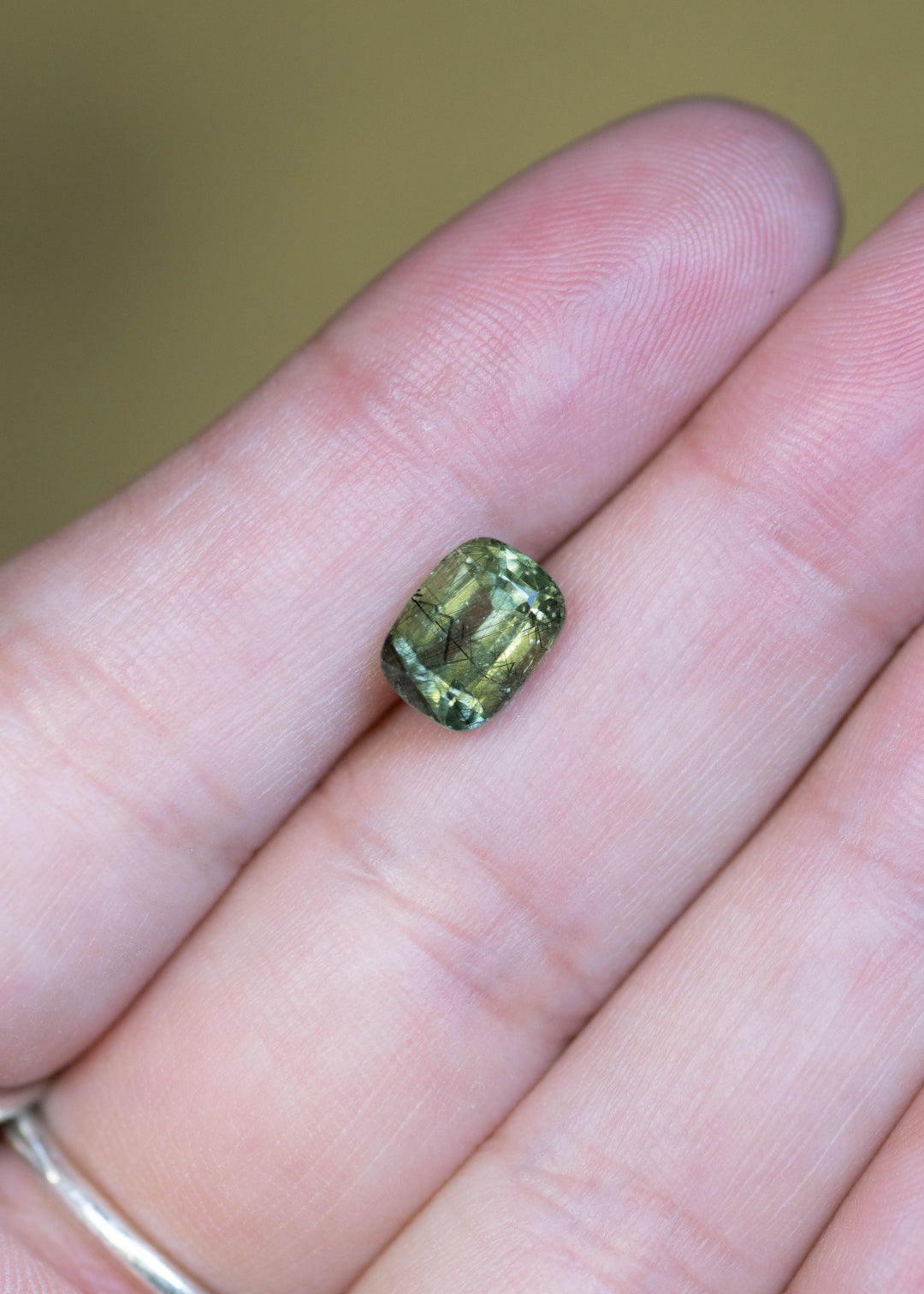 Natural untreated 2.72 carat ombre soft green peridot from Skardu, Pakistan shown on fingertips for scale, displaying its cushion mix cut and subtle green color gradient.