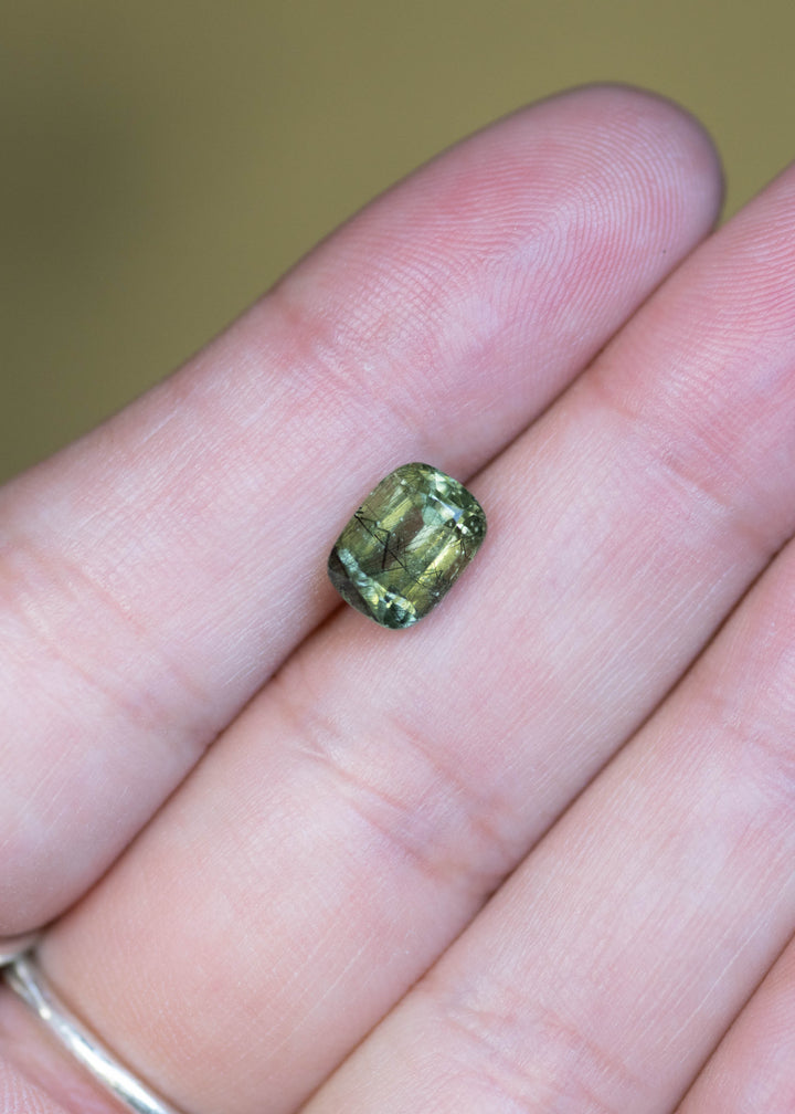 Natural untreated 2.72 carat ombre soft green peridot from Skardu, Pakistan shown on fingertips for scale, displaying its cushion mix cut and subtle green color gradient.