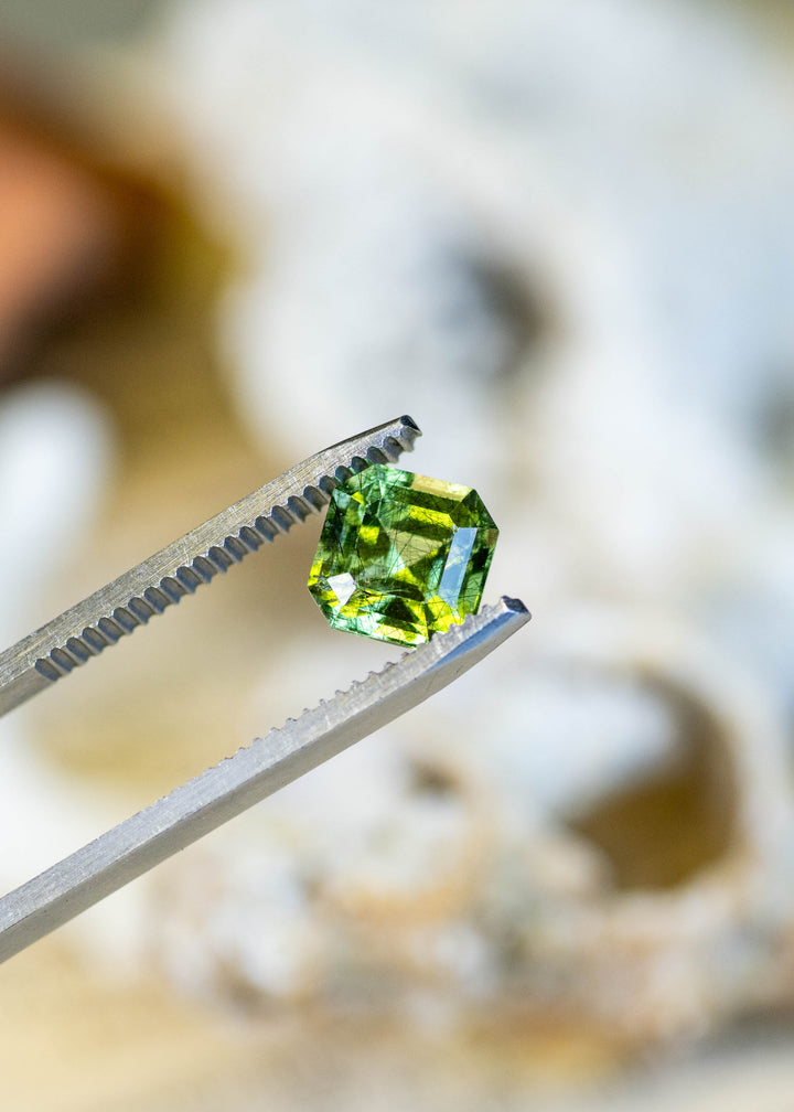 Close-up of a natural Pakistani peridot held in tweezers, highlighting its vivid lime-green color, custom Asscher cut, and intricate natural needle inclusions.