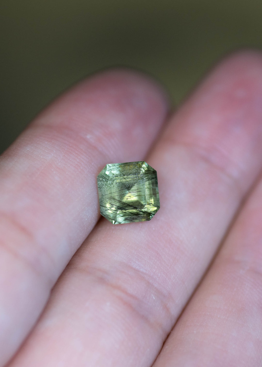 Untreated 2.89 carat Pakistan peridot shown on fingertips for scale, featuring a precision custom Asscher cut and soft sage green coloration.