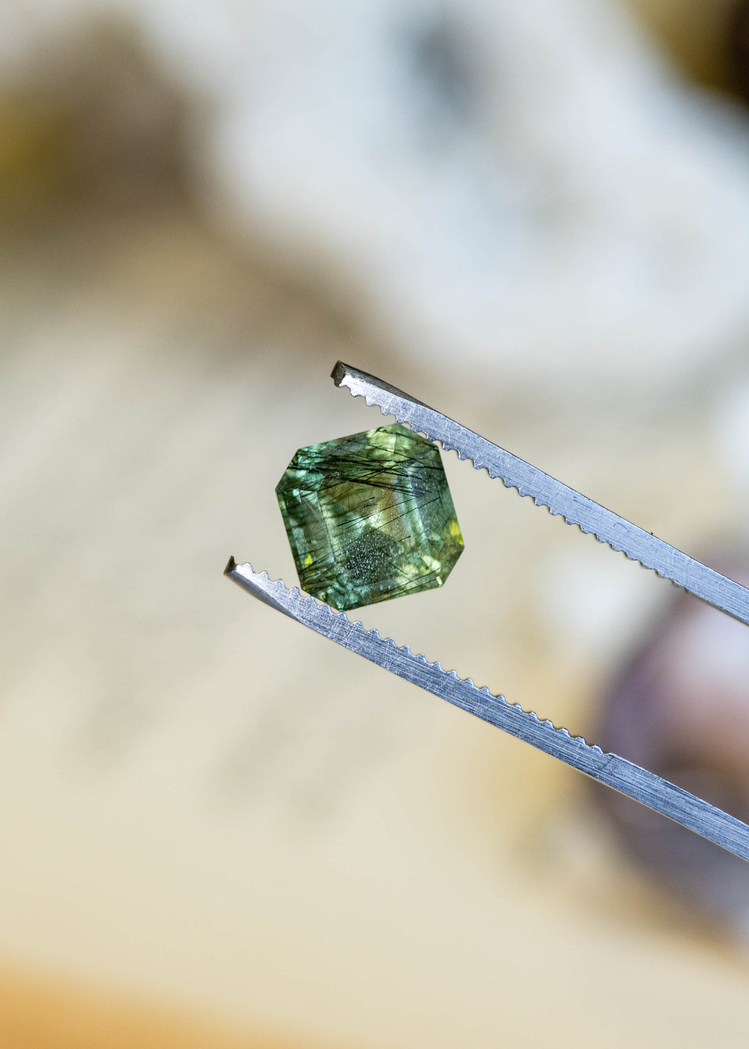 Close-up of a natural Pakistan peridot in tweezers highlighting its custom Asscher cut, soft sage green color, and distinctive internal crystal inclusions.