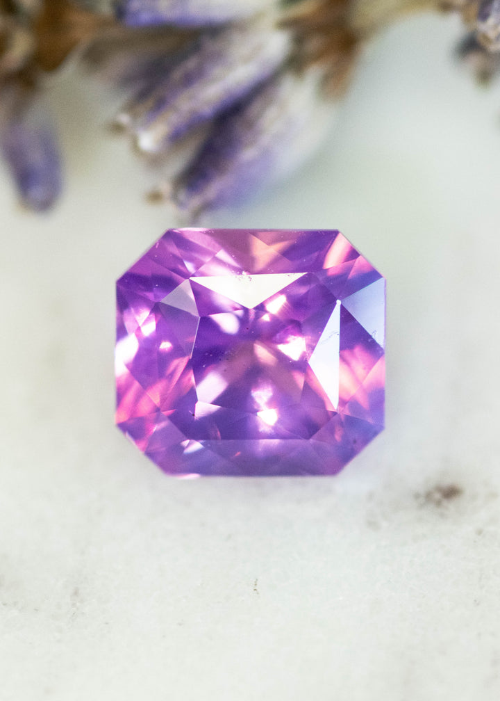 2.00 carat opalescent purple pink sapphire in custom radiant cut resting on a white surface, showing vivid violet body color with rose flashes and soft pastel glow.