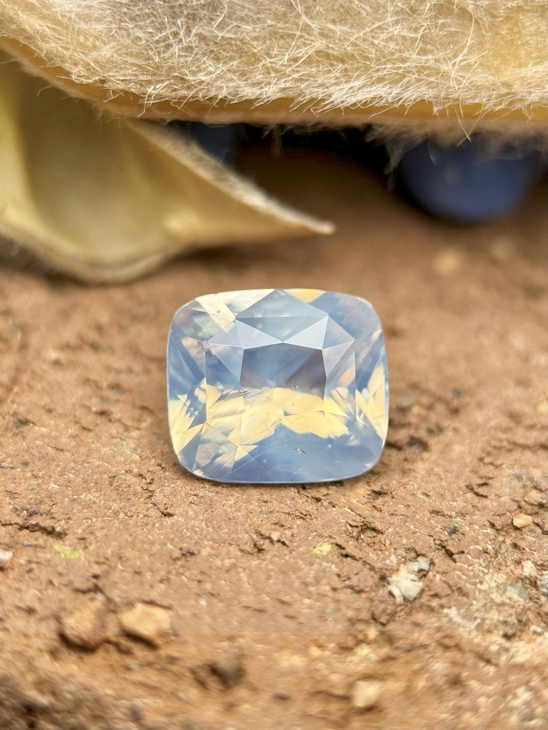 2.00 carat opalescent silver-blue sapphire resting on natural earth-toned surface, showing soft pastel glow and internal light play, untreated cushion cut gem from Ratnapura, Sri Lanka.