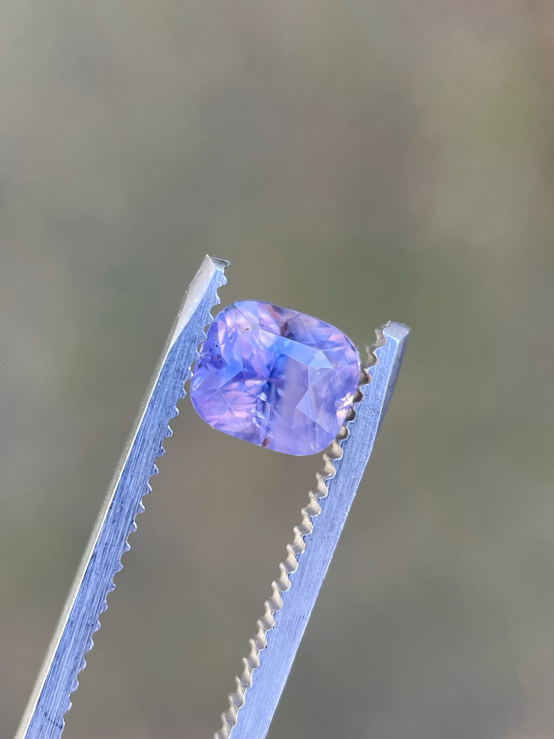 Close-up of a 2.00-carat opalescent pink and violet sapphire held in precision tweezers, displaying its cushion mix cut and intricate facets. Ethically sourced from Sri Lanka, untreated and natural.
