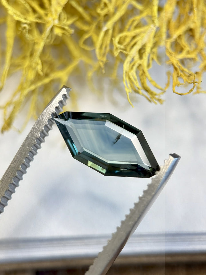 Close-up image of 2.00ct teal-blue sapphire in tweezers showing custom geometric portrait cut.