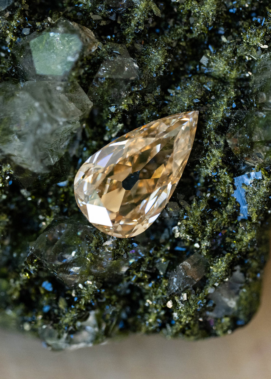 Elongated pear-shaped champagne diamond resting on a dark green crystalline mineral specimen. Broad antique-style facets reflect pale gold, honey and warm amber tones across the stone.