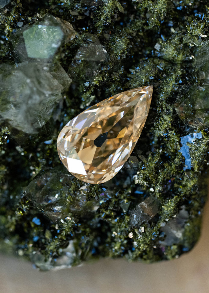Elongated pear-shaped champagne diamond resting on a dark green crystalline mineral specimen. Broad antique-style facets reflect pale gold, honey and warm amber tones across the stone.