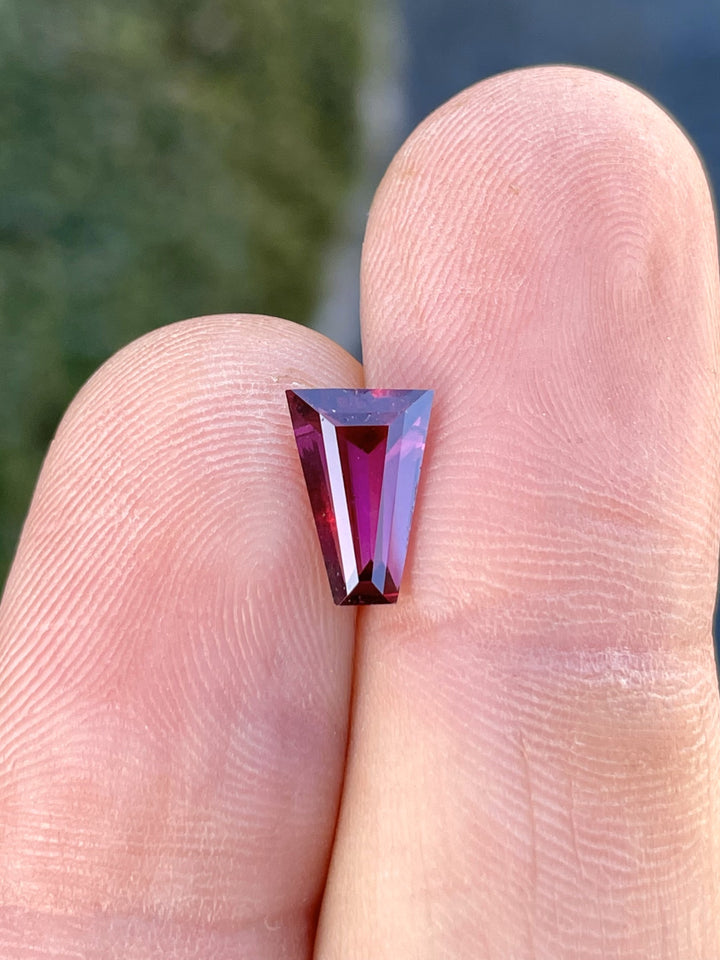 Close-up of a tapered baguette-cut garnet gemstone weighing 2.02 carats, showcasing vibrant red and purple hues, held between fingers.