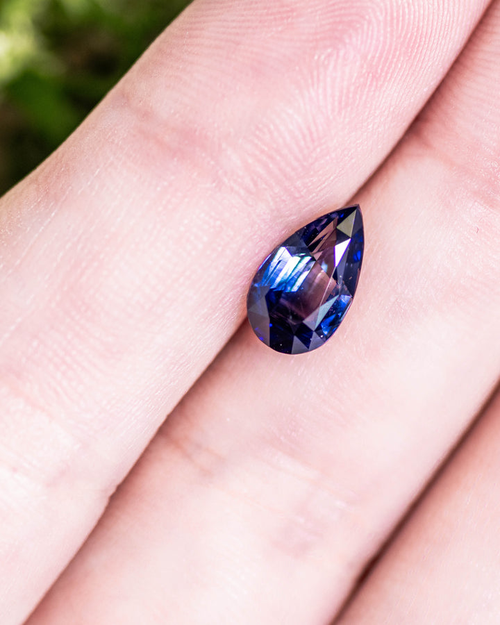 2.02 carat bi-color blue sapphire resting on fingertips, showing deep blue color zoning and sparkle from its pear brilliant cut facets, untreated gemstone from Sakaraha, Madagascar.