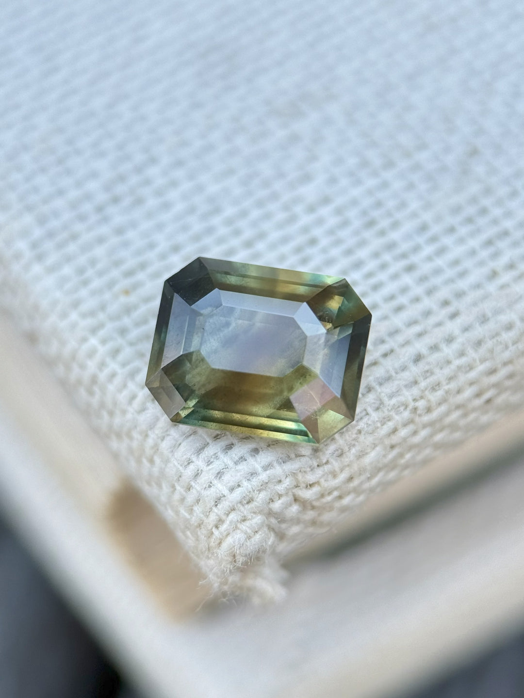 Emerald cut multicolor sapphire with earthy green, brown, and yellow tones resting on textured white fabric – 2.02 carats from Garba Tula, Kenya.
