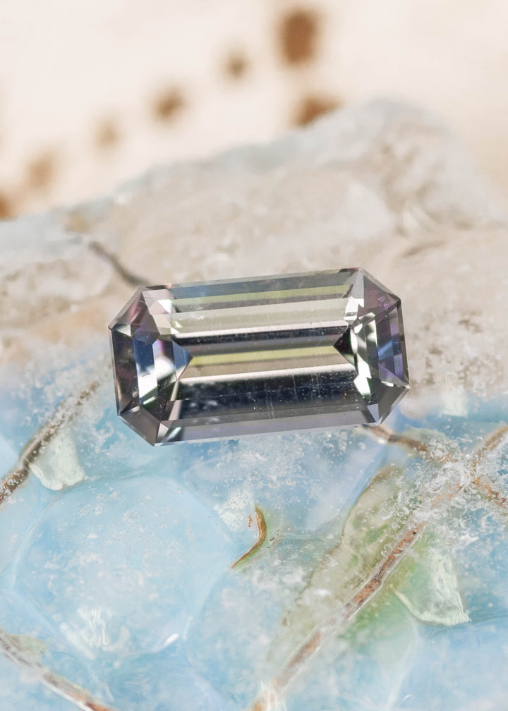 2.02 carat purple green Tanzanite emerald cut placed on display surface, untreated gemstone from Merelani Hills Tanzania highlighting elongated step cut facets, clarity and natural bi color appearance