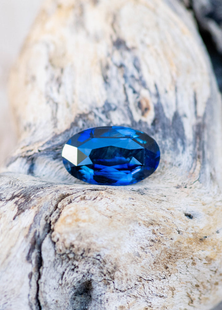 2.02ct deep blue elongated moval cut sapphire from madagascar resting on a textured piece of found driftwood