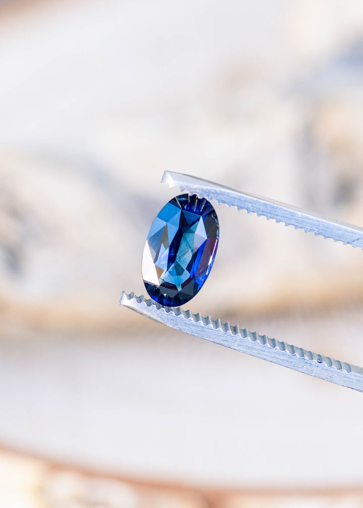 2.02ct deep blue elongated moval cut sapphire from madagascar held in gem tweezers under natural lighting.