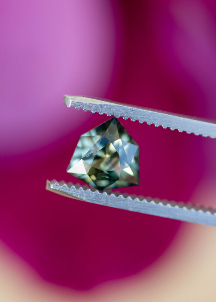 Bluish teal green parti sapphire held in tweezers displaying geometric shield cut, precision faceting, and heated Madagascar origin.