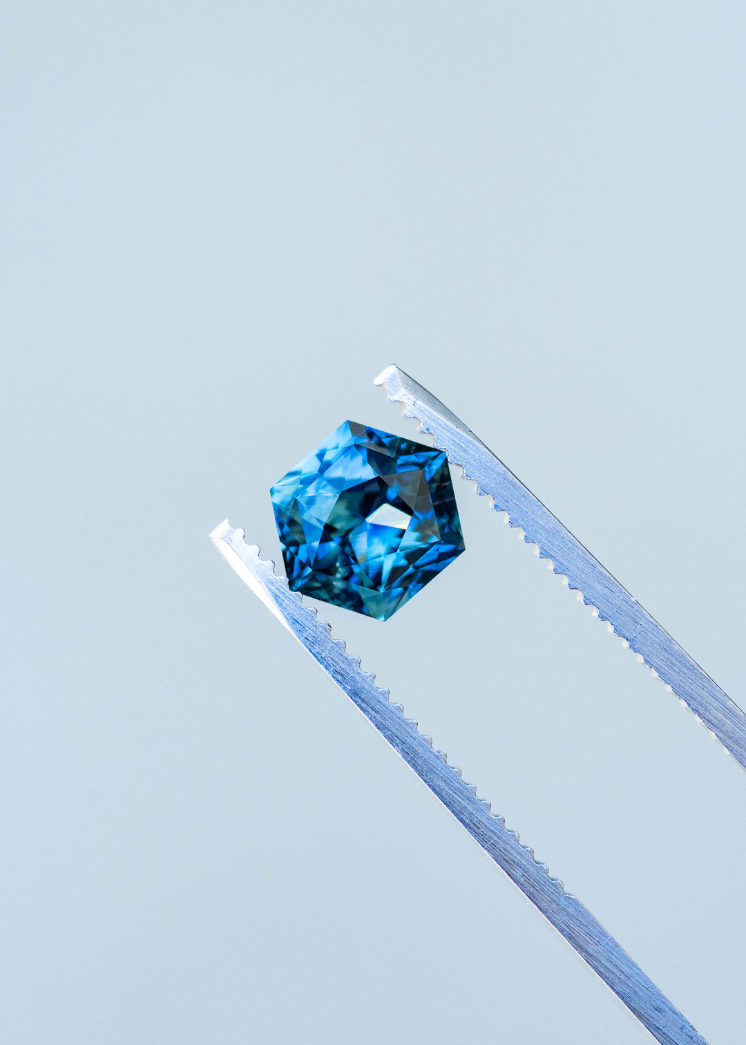 2.03ct blue teal precision hexagonal cut sapphire held in gem tweezers under natural light.