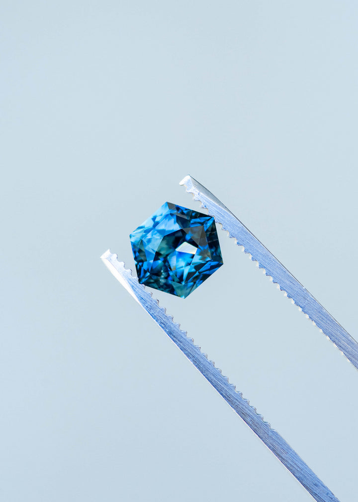 2.03ct blue teal precision hexagonal cut sapphire held in gem tweezers under natural light.
