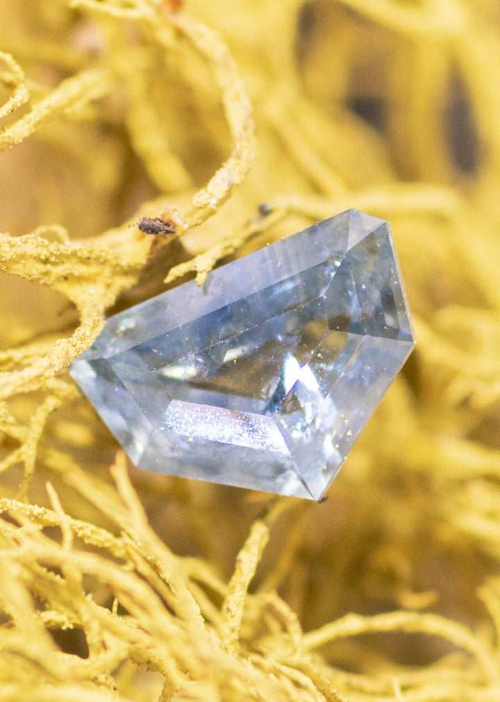 Light blue geometric half moon Montana sapphire resting on golden moss, showing icy blue body color, sharp angular facet junctions, and visible internal inclusions through the center.