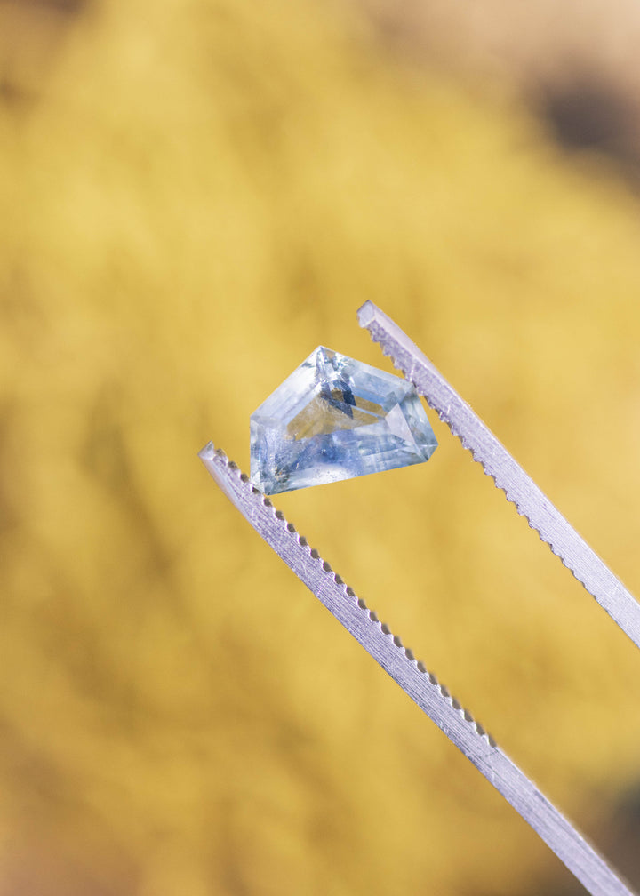2.04ct light blue half moon sapphire held in silver gem tweezers, highlighting the curved outer edge, flat geometric crown facets, and internal feather-like inclusions under natural light.