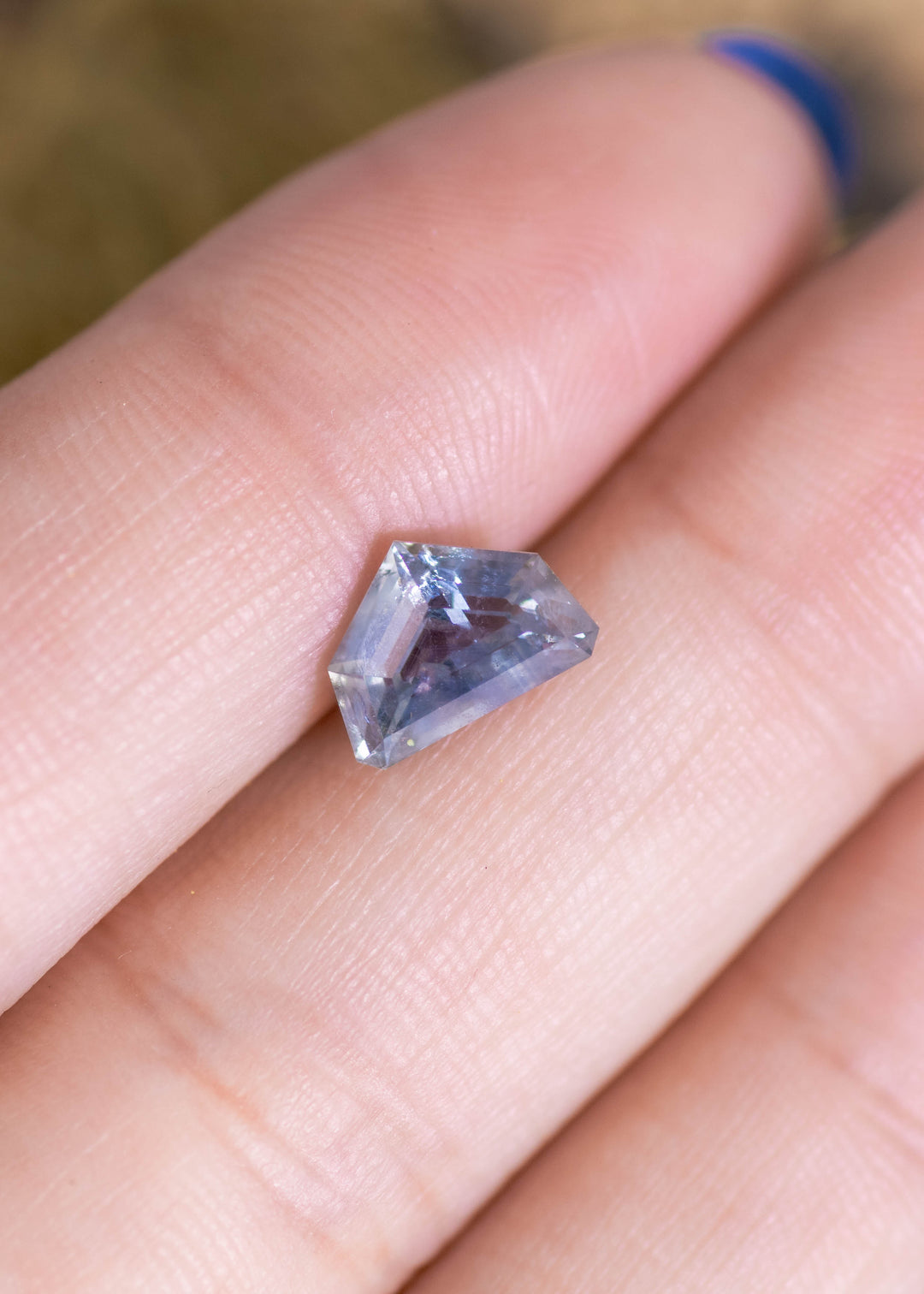 Light blue geometric half moon Montana sapphire on fingertip for scale, displaying its asymmetrical curved outline, pale sky-blue tone, and step-like facet reflections across the surface.