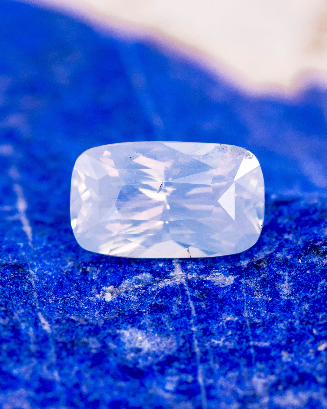 2.05 carat opalescent peach sapphire resting on vivid blue stone background, showcasing pastel reflections and the brilliance of the cushion cut, untreated gemstone from Sakaraha, Madagascar.