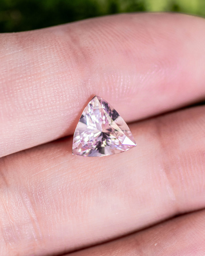 2.06 carat peach and pink sapphire resting on fingertips under daylight, revealing delicate pastel color transitions and sparkle of the trillion cut, untreated gemstone from Sakaraha, Madagascar.