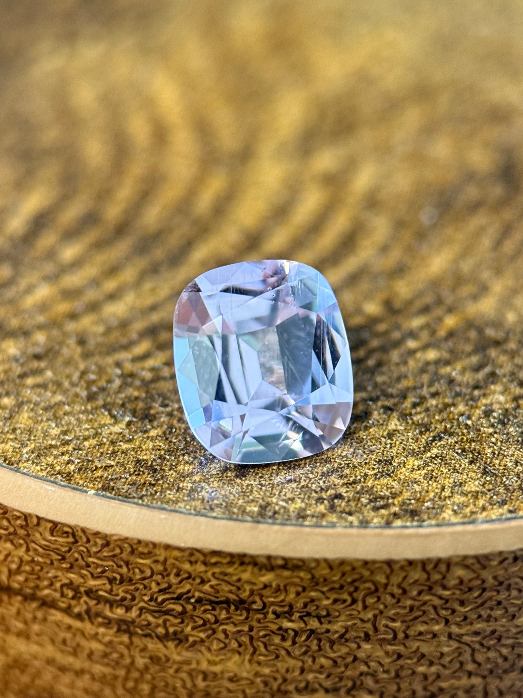 A cushion-cut light blue gemstone with a faceted cushion shape, resting on a textured gold surface.