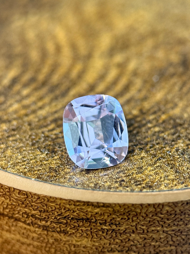A cushion-cut light blue gemstone with a faceted cushion shape, resting on a textured gold surface.