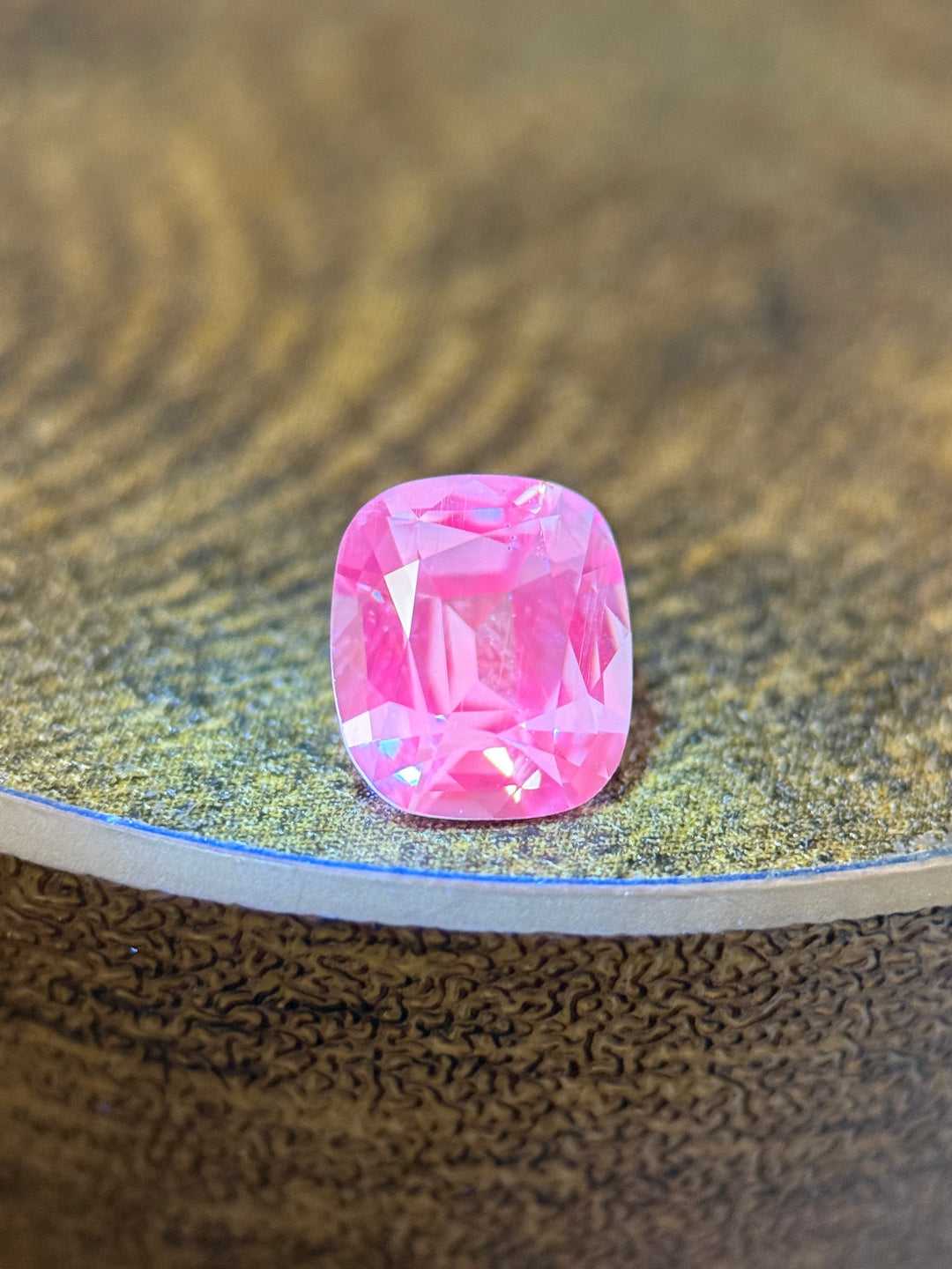 A cushion-cut pink gemstone with faceted edges, resting on a textured metallic surface.