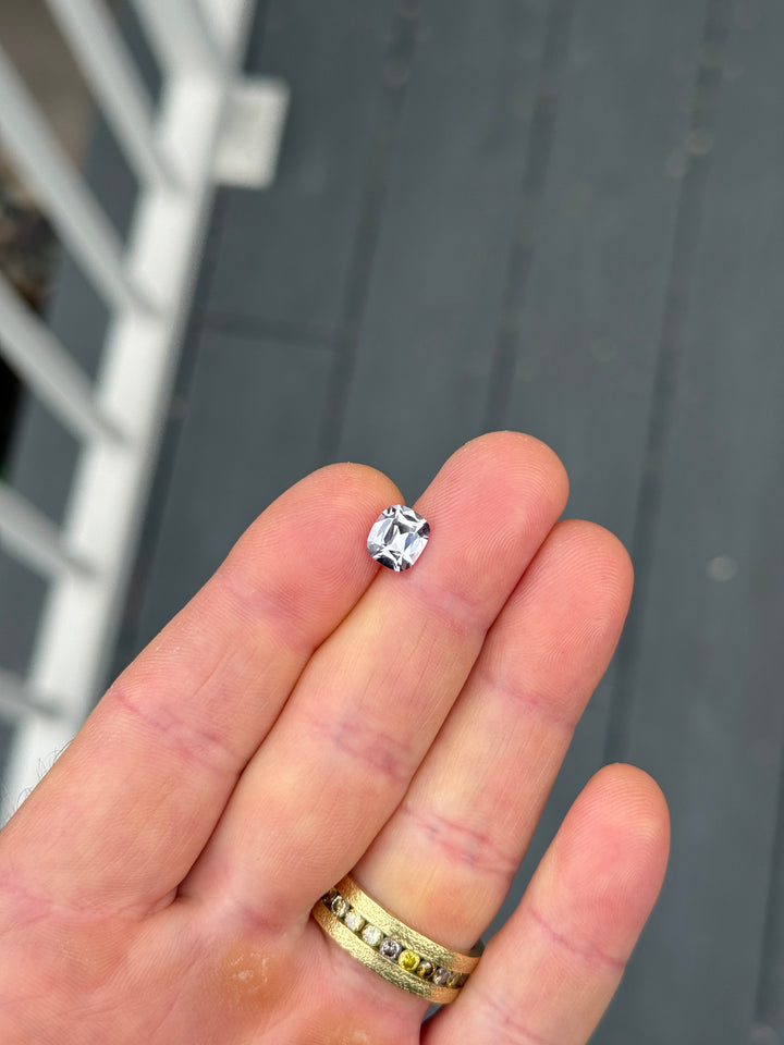A small silver-toned ring with a clear rectangular gemstone held between fingers, with a gold ring on another finger, against a dark gray background.