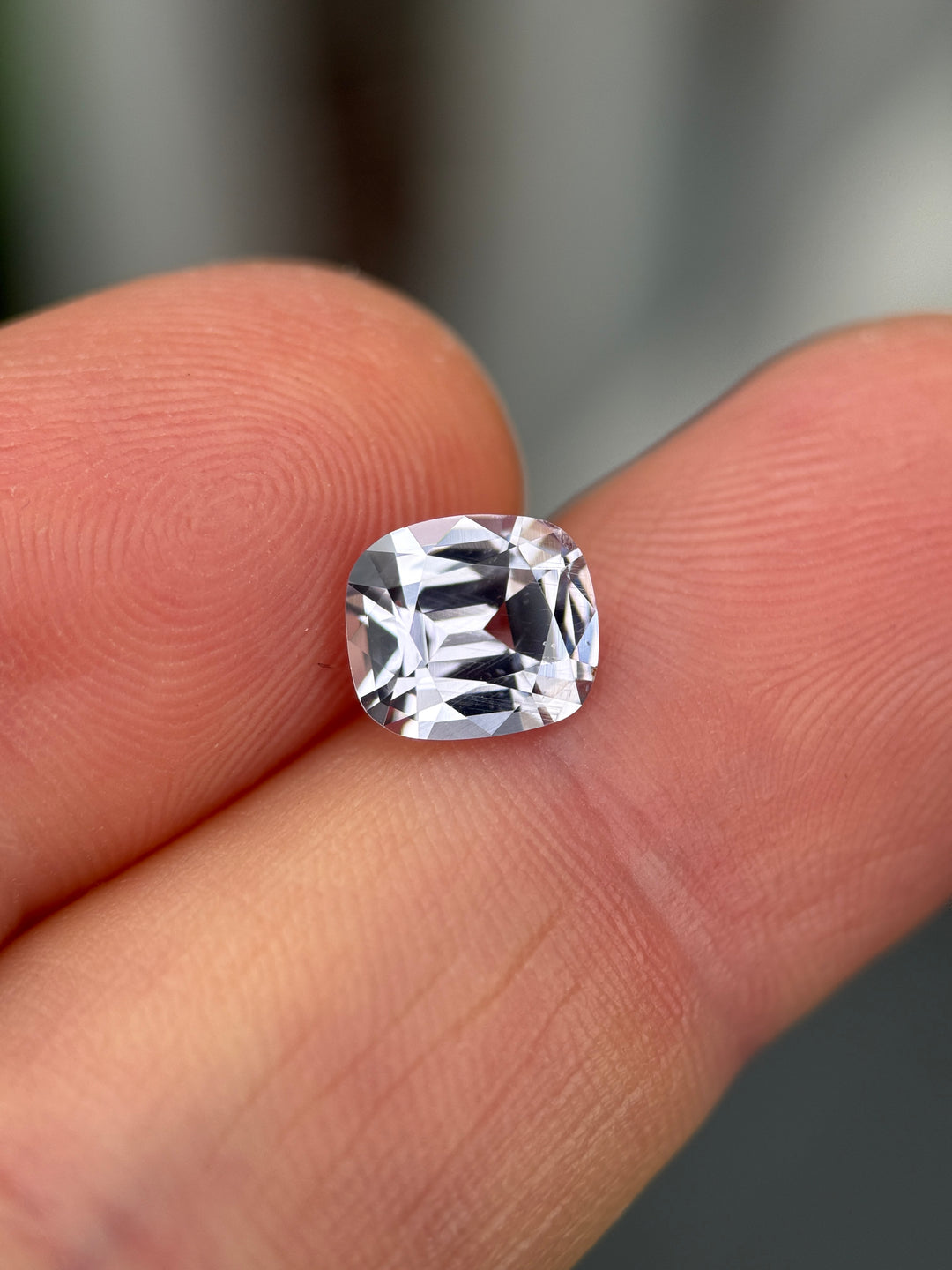 A cushion-cut clear gemstone held between two human fingers to show its size, photographed in close-up.