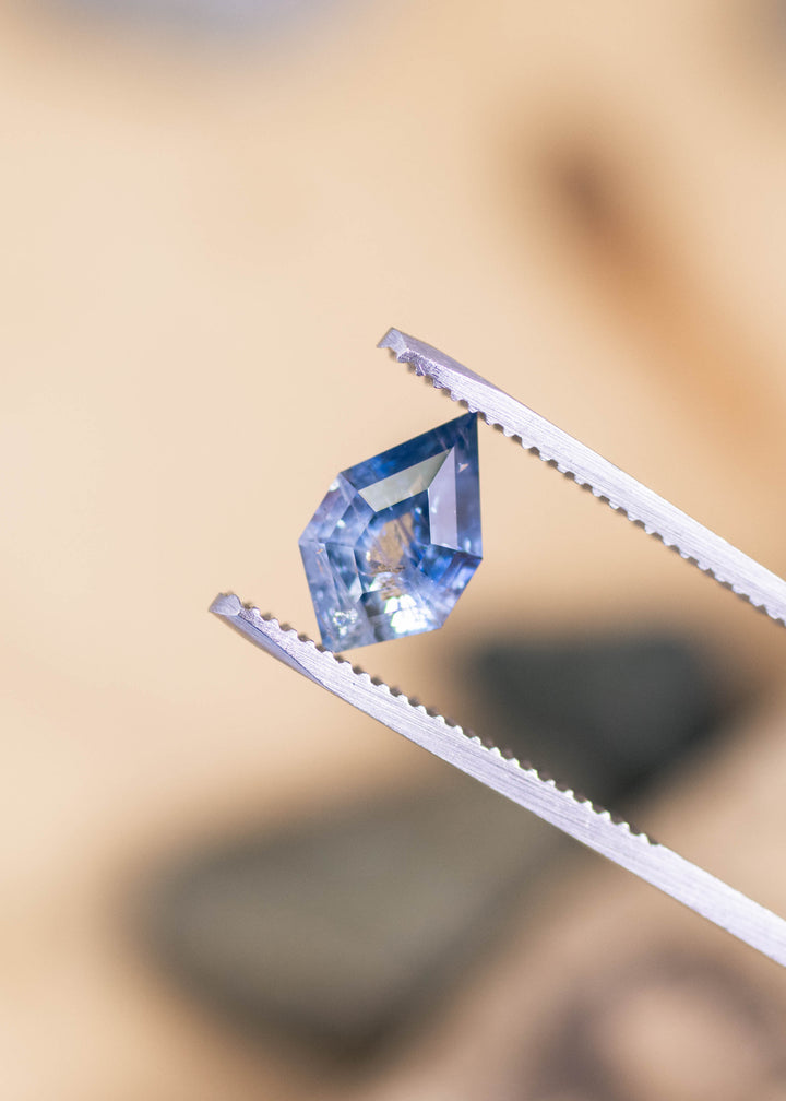 Montana parti sapphire held in metal tweezers, revealing sharp shield-like points and step-style tablet faceting; deep blue flashes appear near one corner while the opposite side shows lighter, milky white transparency with visible internal feather inclusions.