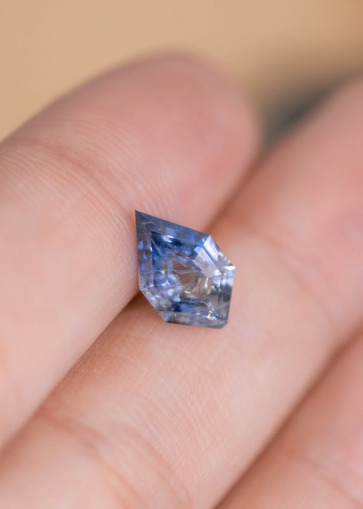 Blue and white heated Montana sapphire balanced on a fingertip; elongated 11mm shield shape shows distinct vertical blue zoning contrasted with pale, icy white sections, with crisp facet edges and visible internal inclusions giving it natural character.