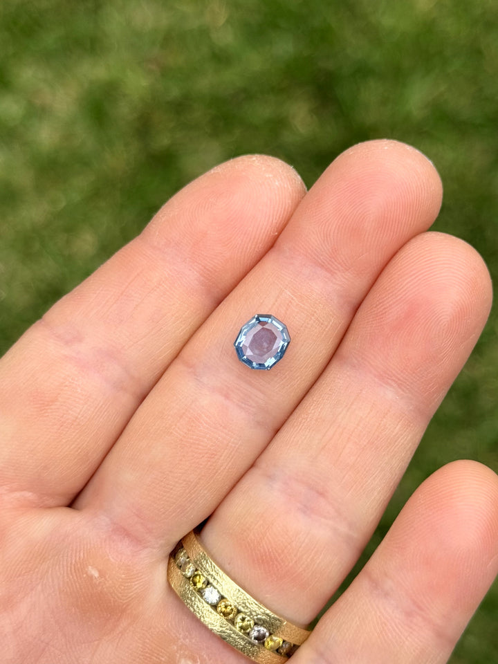 Unheated Montana sapphire showing off its vibrant color-shift blue and violet tones, resting on fingertips with gold ring.