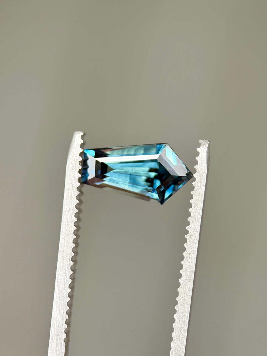 2.11-carat teal blue parti sapphire held in gemstone tweezers, displaying its elongated shield step cut and banded parti color pattern under neutral lighting.