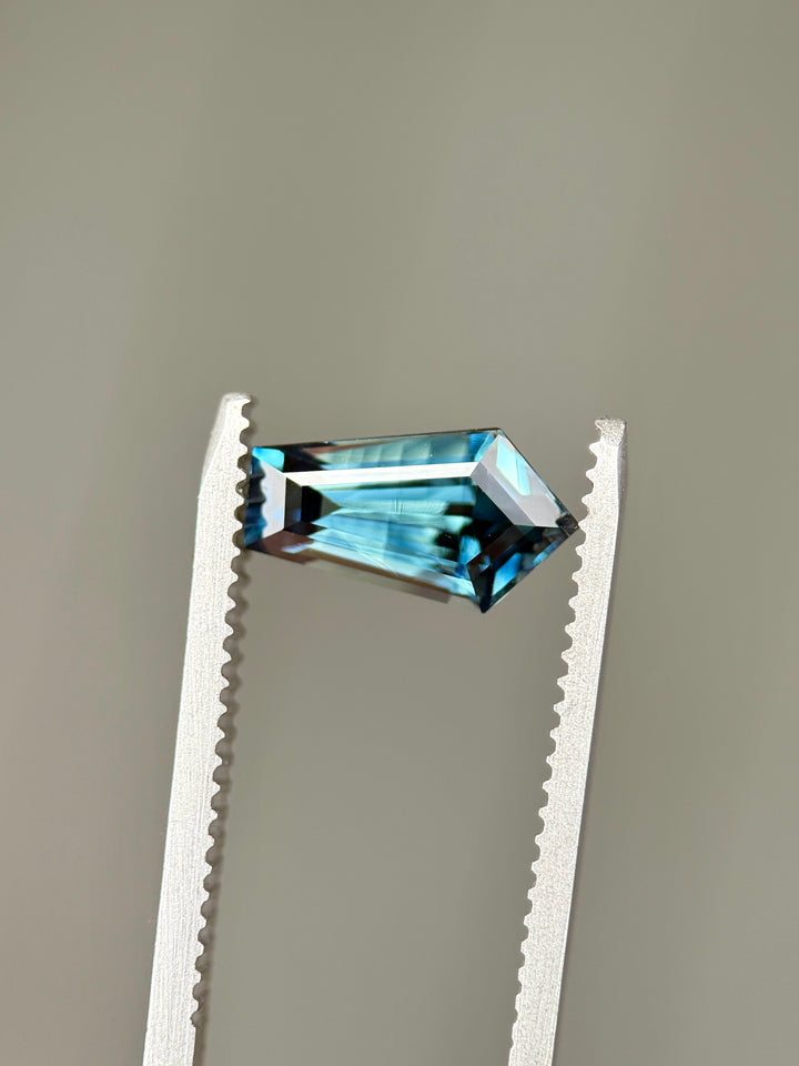 2.11-carat teal blue parti sapphire held in gemstone tweezers, displaying its elongated shield step cut and banded parti color pattern under neutral lighting.