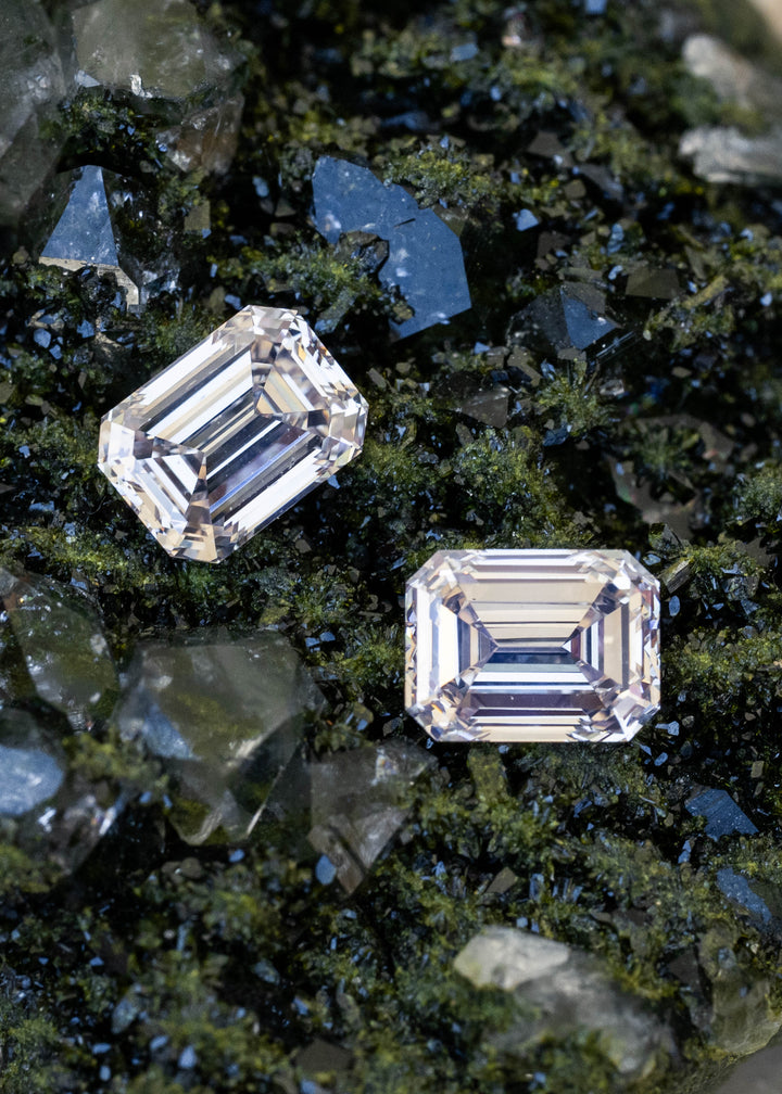 Matched 1.02 and 1.11 carat emerald cut natural diamonds resting separately on dark green mineral crystals, displaying pale rosy champagne color and geometric step-cut reflections.