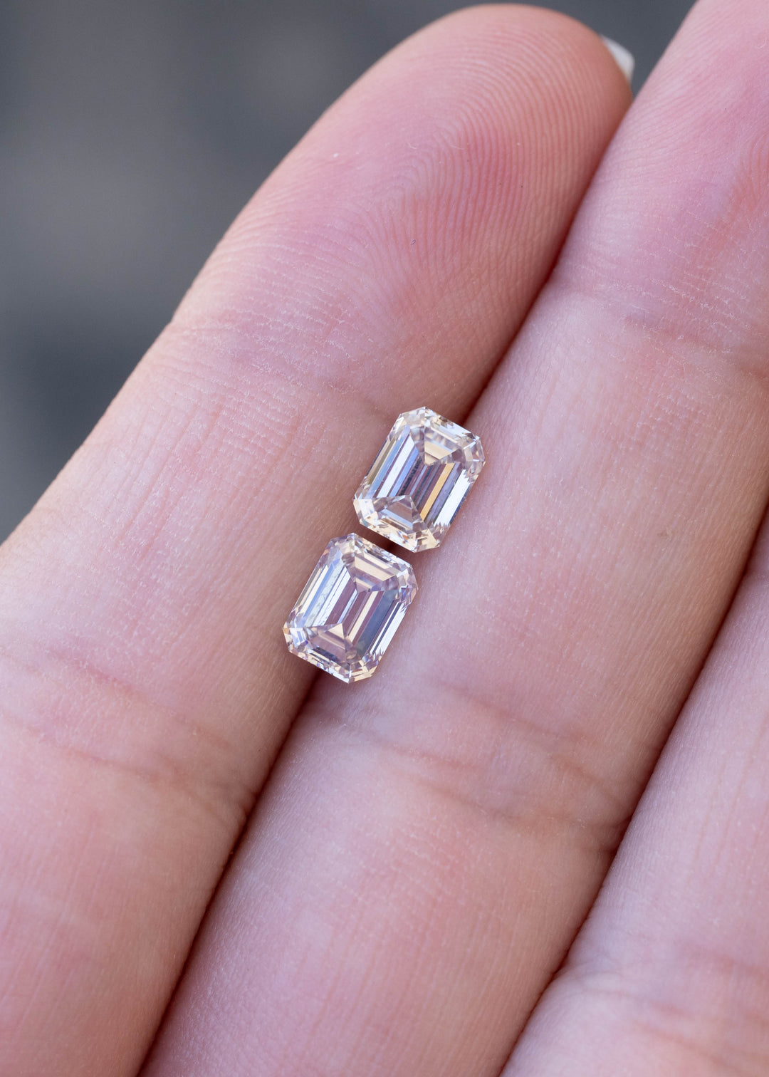 Two light rosy brown emerald cut diamonds arranged vertically on a hand for scale, showing the close match between the 1.02 and 1.11 carat stones.