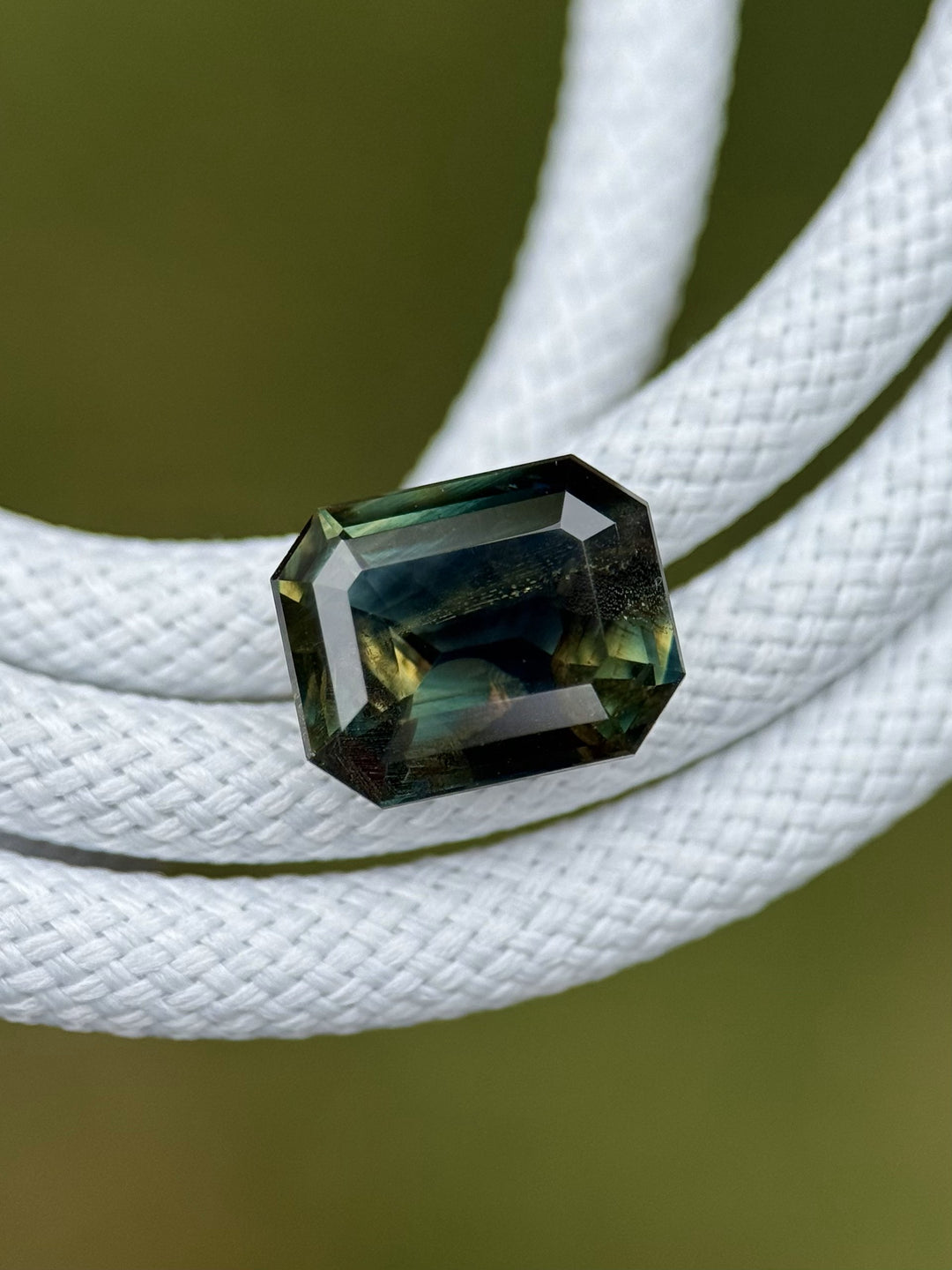 Modified emerald-cut parti sapphire in natural light, lying on white braided rope for contrast.