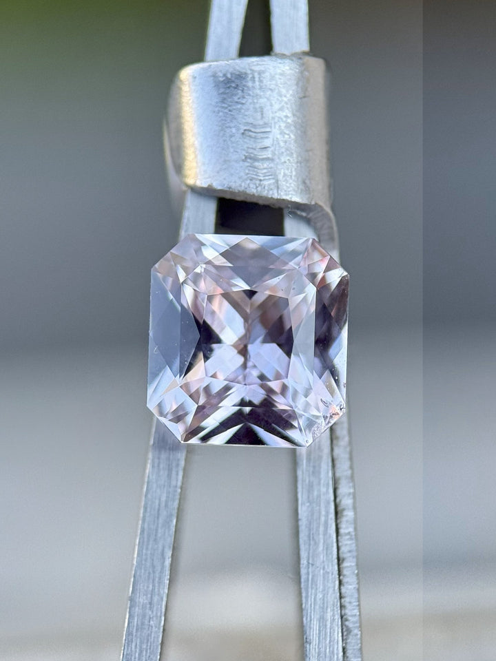 Close-up of a 2.14-carat untreated lavender sapphire from Sakaraha, Madagascar in a custom octagonal radiant cut, displayed in precision gemstone tweezers with a matte metallic grip and a soft green-blurred outdoor background.