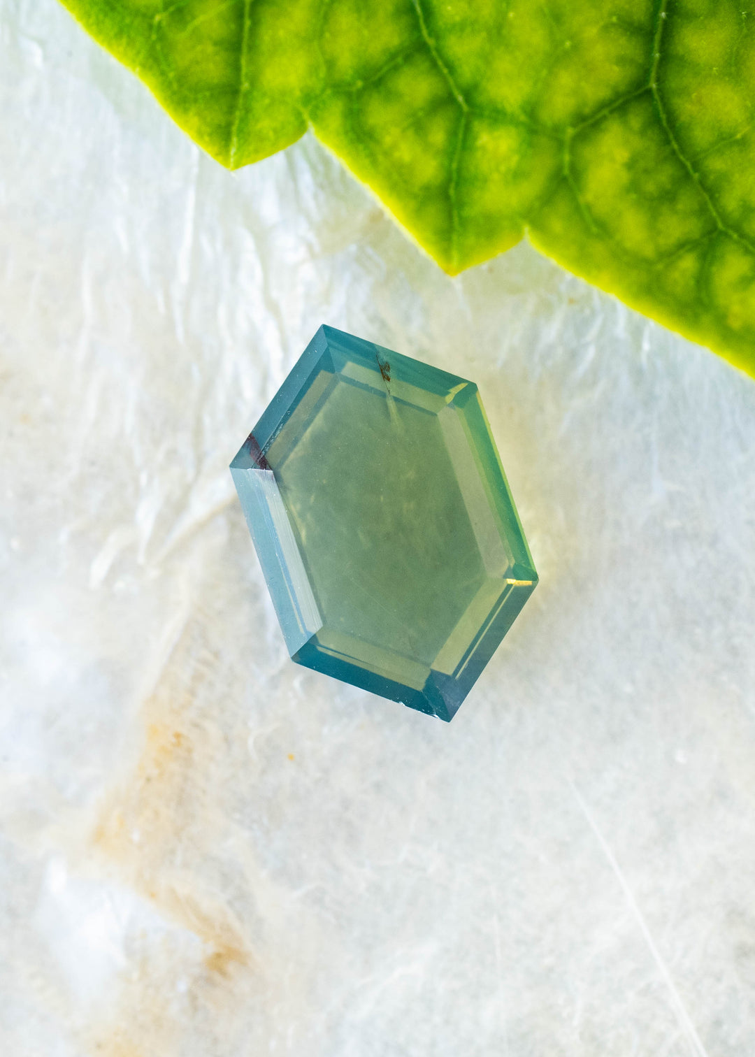 Natural 2.15 carat opalescent teal green sapphire from Gombe, Nigeria, photographed on a pale textured surface beneath a green leaf. Its elongated hexagonal portrait cut reveals a translucent green center framed by deeper teal-blue edges.