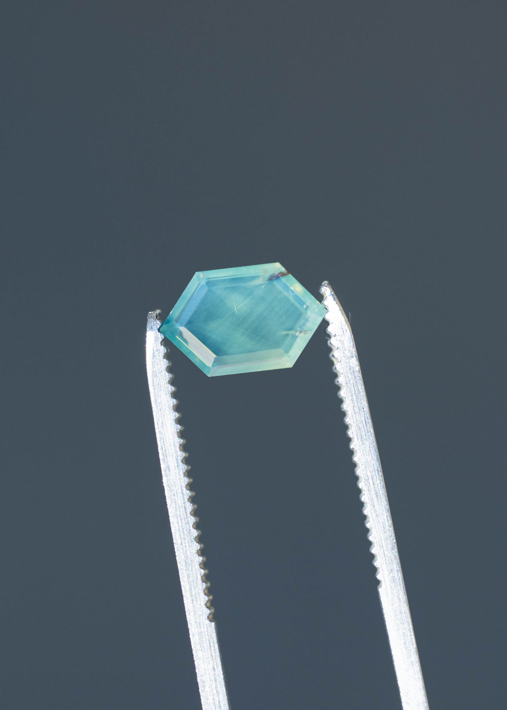 Close-up of an untreated 2.15ct opalescent teal green sapphire held in silver jeweler's tweezers, highlighting its elongated hexagonal outline, broad portrait-cut window and visible internal texture.