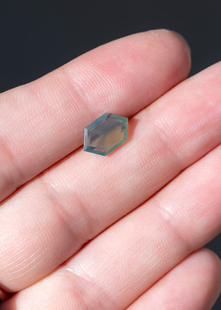 Untreated 2.15 carat opalescent teal green sapphire from Nigeria photographed on fingers for scale. Its elongated hexagonal portrait cut measures 10.70 × 6.67 mm with a shallow 2.56 mm depth.