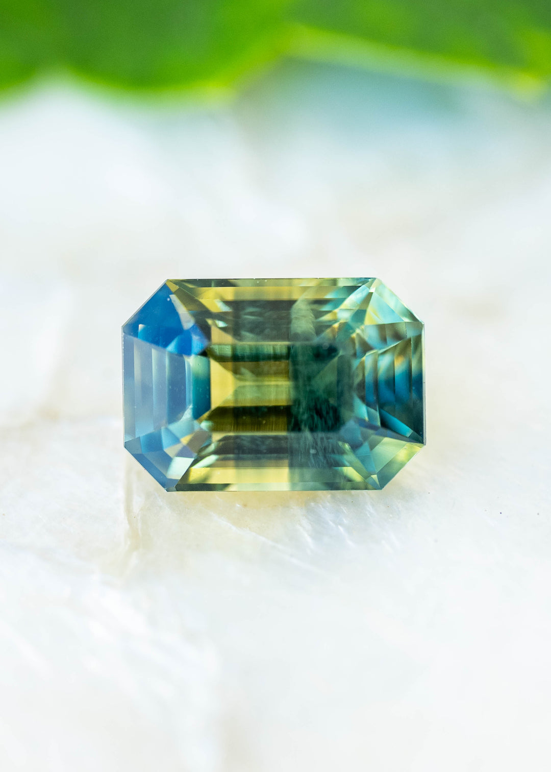 Natural light photograph of a 2.16ct untreated parti sapphire from Diego, Madagascar. Its precision octagonal step cut reveals vivid yellow, teal green and blue color zoning within an elongated geometric silhouette.