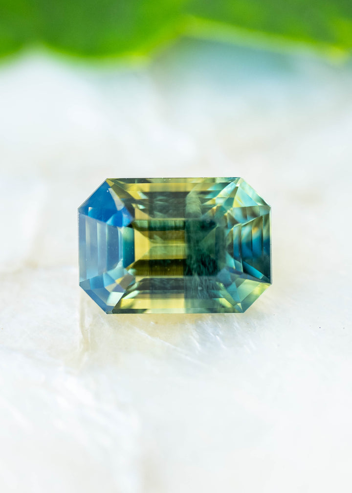 Natural light photograph of a 2.16ct untreated parti sapphire from Diego, Madagascar. Its precision octagonal step cut reveals vivid yellow, teal green and blue color zoning within an elongated geometric silhouette.